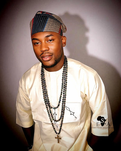 Man wearing a white outfit with our Asooke Cap for Men and multiple necklaces against a plain background