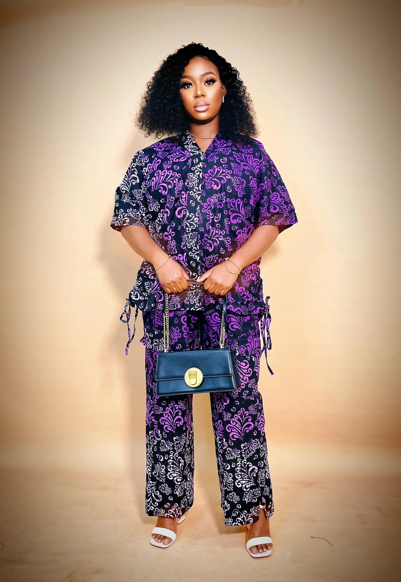 Woman wearing a purple and black African Two Piece Set holding a blue handbag against a beige background